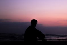 the silhouette of a man sitting on top of a beach next to the ocean at sunset