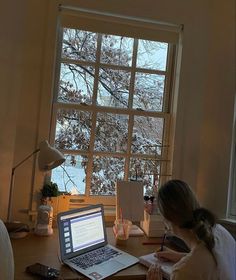 a woman sitting at a desk in front of a window working on her laptop computer