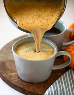 a cup of coffee being poured into two mugs on a cutting board with small pumpkins in the background