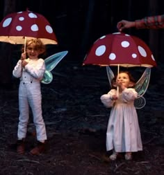 two children holding umbrellas in the woods with white polka dots on them, one girl is wearing a fairy costume