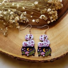 a pair of pink and black earrings sitting on top of a piece of wood next to dried flowers