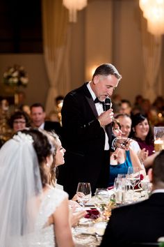 a man in a tuxedo stands at the head of a table while holding a microphone