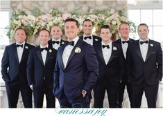 a group of men standing next to each other wearing suits and bow ties in front of a floral wall