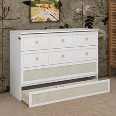 a white dresser with drawers in front of a wallpapered background and painting on the wall