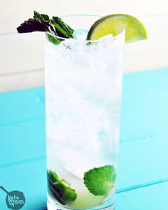 a glass filled with ice and lime on top of a blue table next to a slice of lime