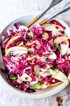 a white bowl filled with red cabbage, apples and walnuts next to two serving utensils