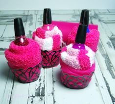 three pink cupcakes with black tops are sitting on a white wooden table and one has two bottles in the shape of hearts