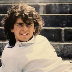 a young man with long hair sitting on steps wearing a white shirt and smiling at the camera