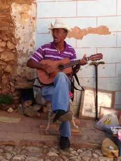 a man sitting on a chair playing a guitar in front of a brick wall with other items around him