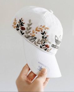 a person holding up a white hat with embroidered flowers on the front and side of it