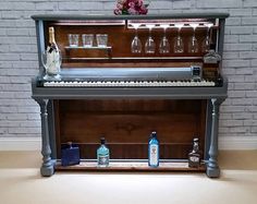 an upright piano with liquor bottles and glasses on the top shelf, in front of a brick wall