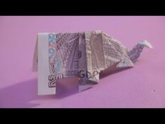 an origami bird made out of money sitting on a pink surface with purple background