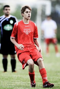 a young man kicking a soccer ball on top of a green field with other people in the background