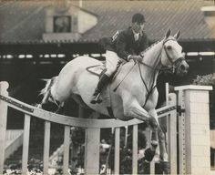 a man riding on the back of a white horse jumping over a wooden fence in front of a building