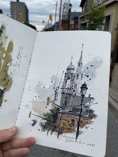 a person holding up an open book with watercolors on it in front of some buildings