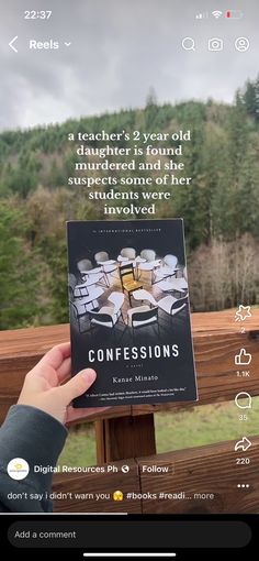 someone holding up a book in front of a wooden fence with trees and mountains behind it