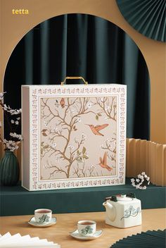 a white box with birds on it sitting next to tea cups and saucers in front of a window