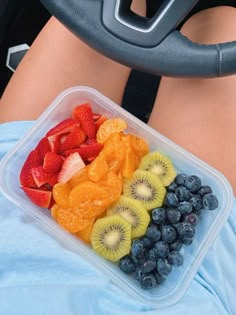 a plastic container filled with assorted fruit on top of a blue blanket next to a steering wheel