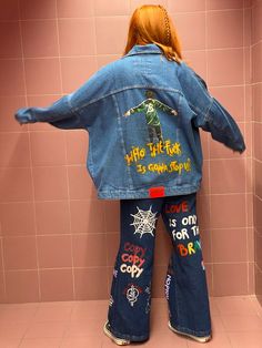 a woman standing in front of a pink tiled wall wearing a jean jacket with writing on it