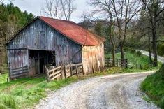 an old barn sits on the side of a dirt road next to a wooded area