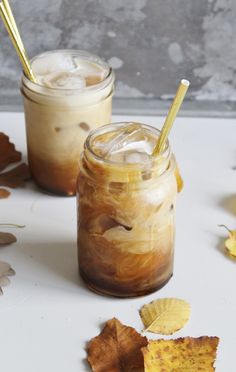 two jars filled with iced coffee next to autumn leaves on a white counter top and one has gold straws in it