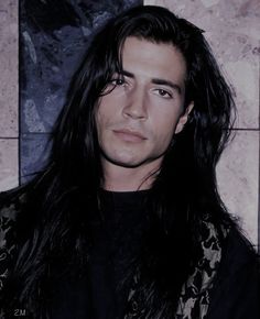 a man with long black hair standing in front of a tile wall and looking at the camera