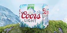 two cans of coors light sitting on top of a moss covered hill with mountains in the background