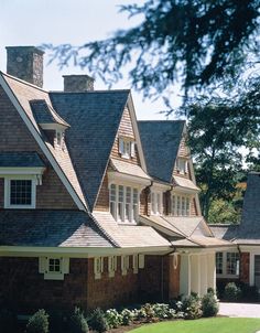 a large brown brick house with white trim and dormers on the roof, surrounded by greenery