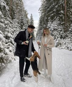 a man and woman standing next to a dog in the snow holding wine bottles while they look at each other