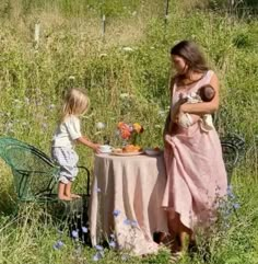 a woman holding a baby next to a table with food on it in a field