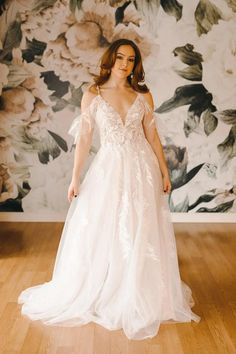 a woman standing in front of a floral wallpaper wearing a white wedding dress with sheer sleeves