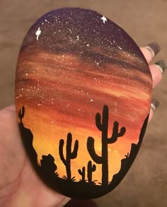 a hand holding up a painted rock with cactus silhouettes on it and stars in the sky