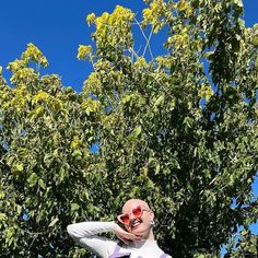 a man wearing sunglasses and a white shirt is standing in front of a tree with his hands behind his head