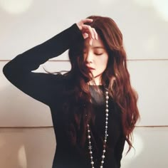 a woman with long brown hair wearing a black top and pearls on her necklace, standing in front of a white wall