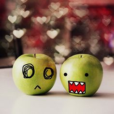 two green apples with faces drawn on them sitting next to each other, one has its mouth open and the other has it's eyes closed