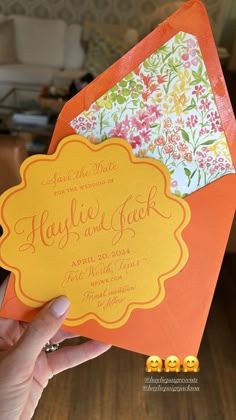 a person holding up an orange and yellow wedding card with flowers on the envelopes