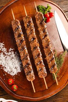 three skewers of meat sitting on top of a wooden plate next to tomatoes and parmesan cheese