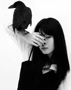 a woman with long black hair holding a bird on her shoulder while standing in front of a white wall