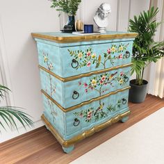 a blue dresser with flowers painted on the front and gold trimmings, next to a potted plant