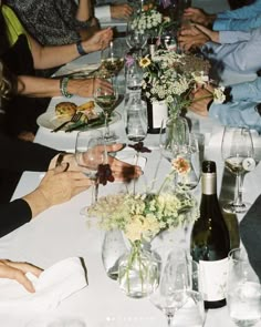 a group of people sitting at a dinner table with wine glasses and flowers in vases