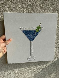 a hand holding up a small canvas with a blue martini on it, in front of a gray wall