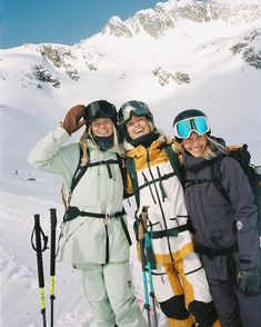 three skiers pose for a photo in front of a snowy mountain range with their ski equipment