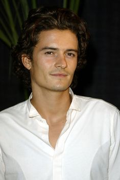 a man with curly hair wearing a white shirt and looking at the camera while standing in front of a plant