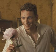 a man holding a pink flower in his right hand and smiling at the camera,