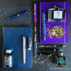 the contents of a fountain pen, notebook and other items on a black surface with gold confetti sprinkles