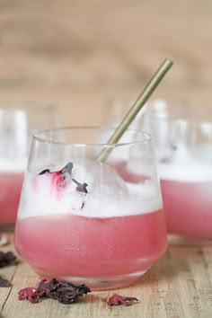 two glasses filled with pink liquid and ice on top of a wooden table next to dried flowers