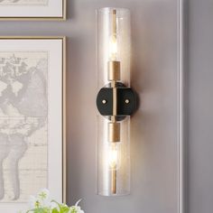 a wall mounted light with two lights on it next to a vase and framed pictures