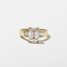 an emerald cut diamond ring with three baguets in yellow gold, on a white background