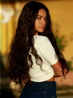 a woman with long dark hair standing in front of a yellow building and looking off into the distance