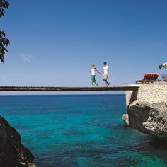 two people walking across a bridge over the ocean on a sunny day with clear blue water